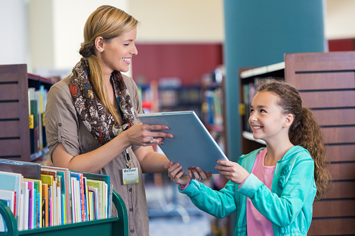 Mid adult Caucasian woman is smiling while helping elementary age little girl select a book in modern public library. Studenet is smiling and looking up at librarian while taking book to borrow. Librarian is pushing green rolling cart full of colorful childrens' books.