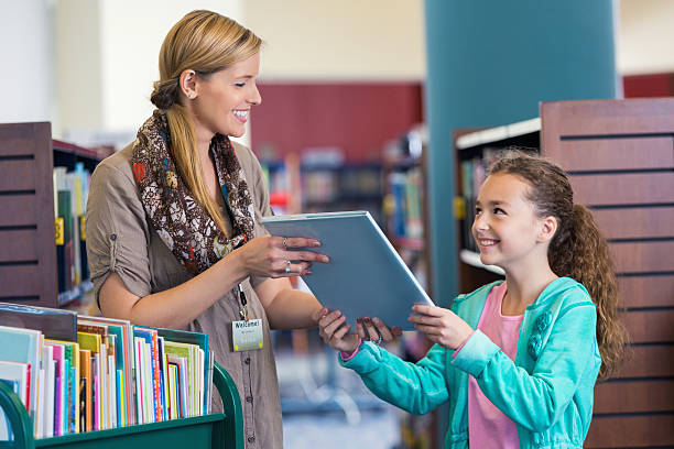 Librarian helping elementary age little girl with book selection Mid adult Caucasian woman is smiling while helping elementary age little girl select a book in modern public library. Studenet is smiling and looking up at librarian while taking book to borrow. Librarian is pushing green rolling cart full of colorful childrens' books. librarian stock pictures, royalty-free photos & images