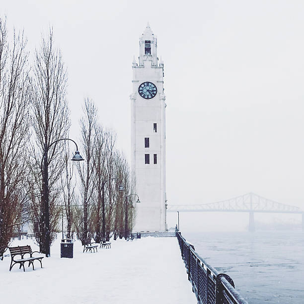 This is a square, color, royalty free stock photograph of a snow covered scene in urban Old Montreal, Quebec Canada. A tall clock tower stands in front of the Pont Jacques Cartier Bridge. Photographed with a Samsung Galaxy S5.