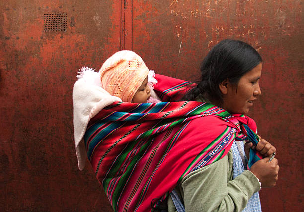La Paz, Bolivia - April 19, 2010: An indigenous Bolivian woman walking next to a red wall in La Paz carrying a baby on her back; the baby is wrapped in a traditional red striped blanket.  