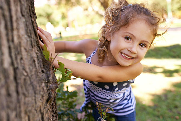 They won't find me here Portrait of a little girl hiding behind a tree in a park 4 year old girl stock pictures, royalty-free photos & images