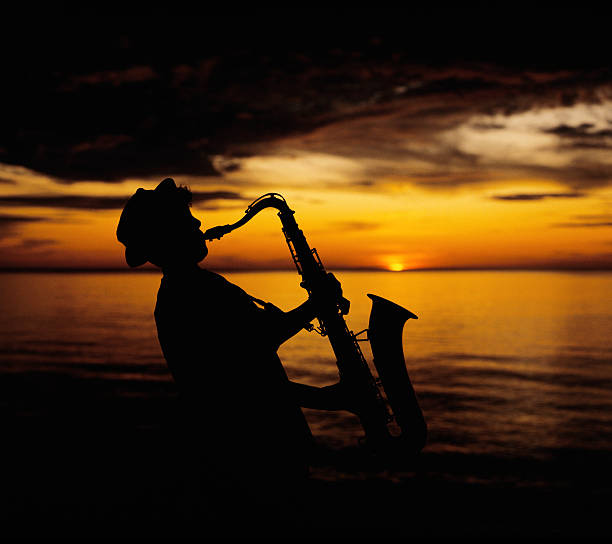 A young teen boy wearing a funky hat plays his saxophone on the beach at sunset.  The young musician is silhouetted against the yellow sky with the setting sun positioned over the end of the saxophone.  Scanned 6x7 film.