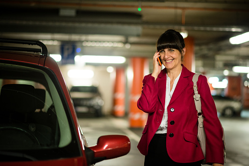 Smiling mature business woman wearing red jacket calling mobile phone while standing at her car