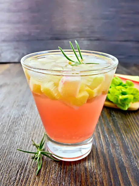 Lemonade with rhubarb and rosemary in a glass, the stems and leaves of rhubarb on a wooden boards background
