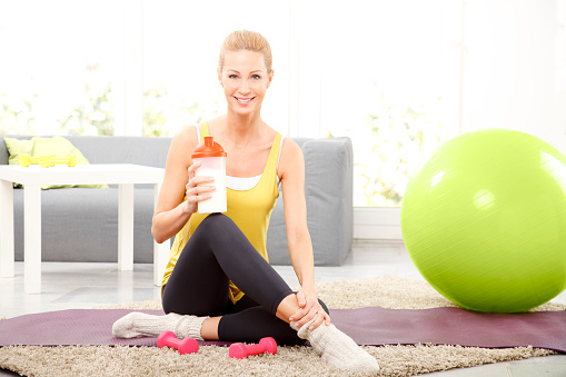 Portrait of middle age woman holding in her hand a shaker while sitting on a yoga mat at home and relaxing after fitness workout.