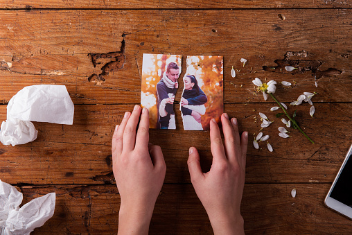 Unrecognizable woman holding torn picture of couple in love. Ended relationship. Crying.Valentines day composition. Studio shot on brown wooden background.