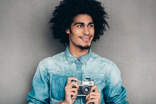 Handsome young African man holding retro styled camera and looking away with smile while standing against grey background