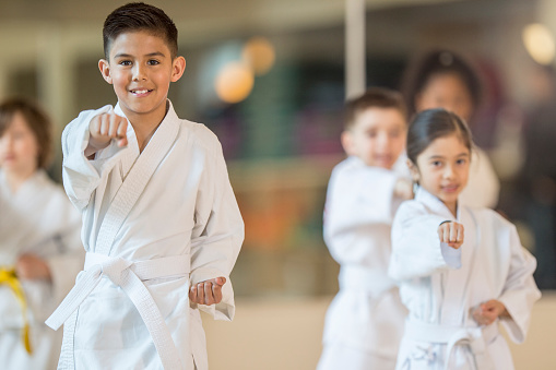 A multi-ethnic group of elementary age children are standing together in formation during their taekwando class. One boy is smiling while looking at the camera.