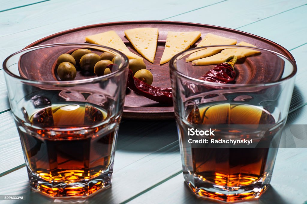 Two whiskey glasses on a wooden background. Two whiskey glasses and a plate of olives, cheese and pepper on a blue wooden background. Cognac - Brandy Stock Photo