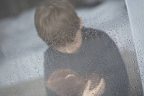 The little boy is hugging his teddy bear. Selective focus.  Standing behind wet from rain window glass. Rainy Day. Loneliness and waiting concept