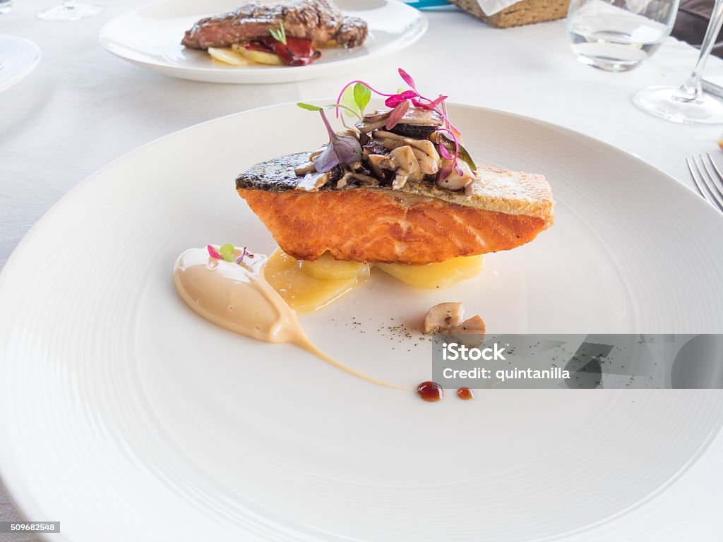 salmon modern dish elegant white circular plate with salmon piece, mayonnaise, potato and mushroom and color leaves on white tablecloth in restaurant Gourmet Stock Photo