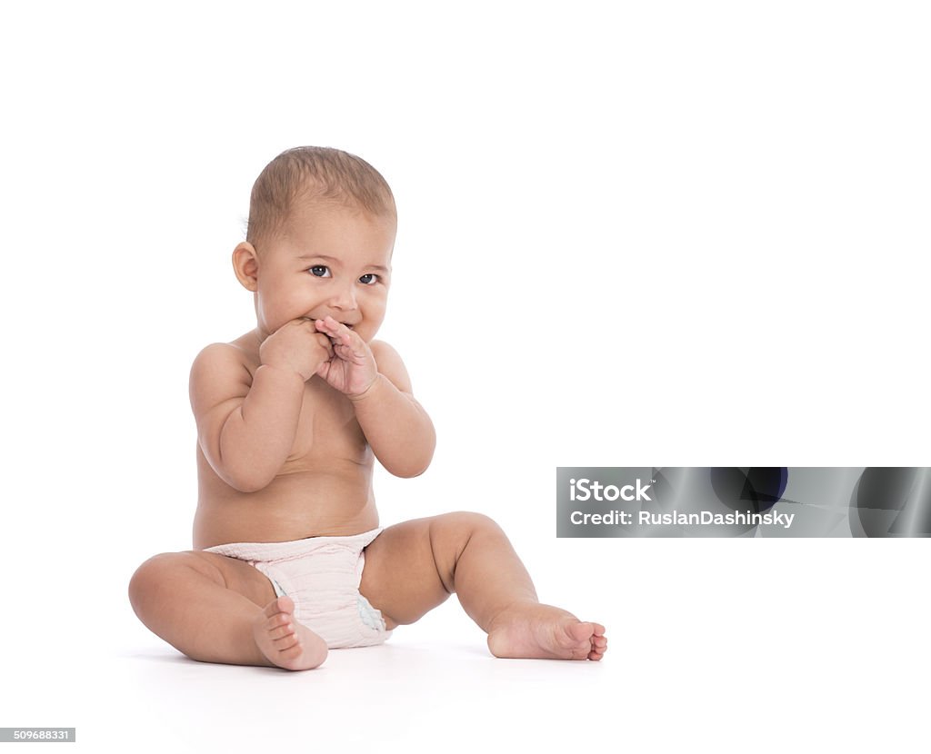Portrait of shy baby with both hands in mouth. Studio shot of adorable 7 month old baby sitting on floor  with two hands in mouth, wearing dipper. Isolated on white background, cut out image, with copy space. White Background Stock Photo