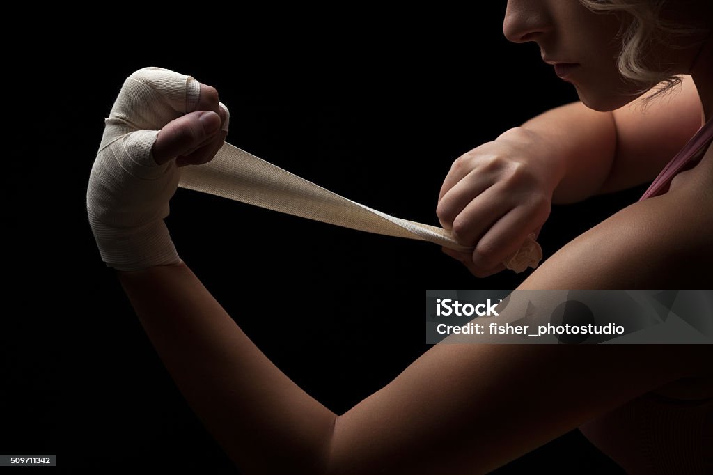 Ready for fight. Beautiful and fit female fighter getting prepared for the fight or training, wrapping her hands with bandage tape against dark background. Active Lifestyle Stock Photo