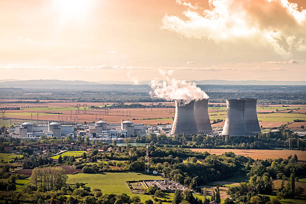 Nuclear power station aerial view in countryside landscape during sunset Horizontal composition photography of french nuclear power station with four steaming cooling towers in countryside plain with smoke cloud (water condensation). Image taken from high angle view, aerial view, in Bugey, in Ain on the border of Isere department, Rhone-Alpes region in France (Europe). The nuclear power station is located in the middle of a plain landscape in France, near Lyon city. This picture was taken during a bright orange sunset in autumn season with green and brown meadow and field. ain france photos stock pictures, royalty-free photos & images