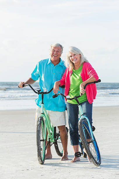 matura coppia biciclette equitazione sulla spiaggia - mature adult bicycle senior adult heterosexual couple foto e immagini stock