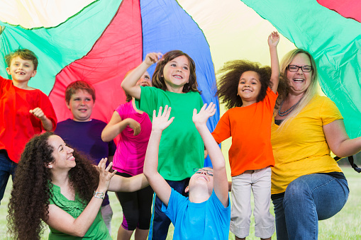 A multi-ethnic group of children with two camp counselors playing under a colorful parachute at the park.  They are dancing and having fun at summer camp.
