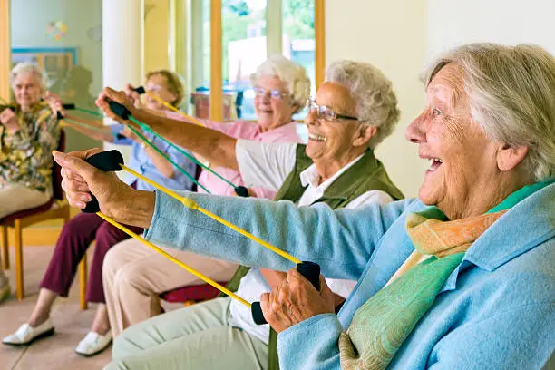 Large group of happy enthusiastic elderly ladies exercising in a gym sitting in chairs doing stretching exercises with rubber bands.