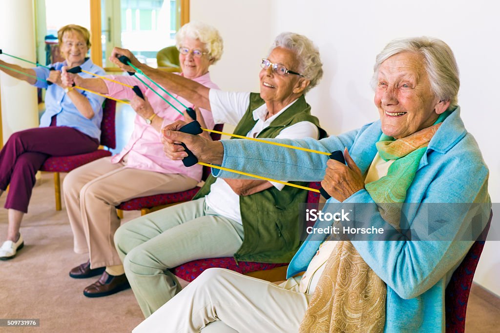Cheerful senior women exercising their arms. Group of four smiling senior women toning their arms with elastic strengthening bands while seated in fitness class. Senior Adult Stock Photo
