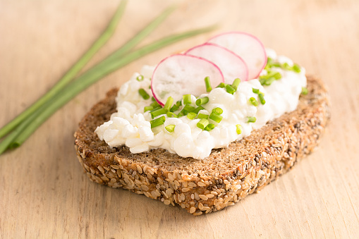 A slice of bread topped with granular cream cheese, radishes and chives lies on a wooden board.