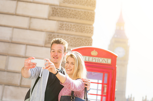 London, happy young couple taking a selfie with a red phone booth and Big Ben on background. They are on holidays for valentines day or they are enjoying their honeymoon.