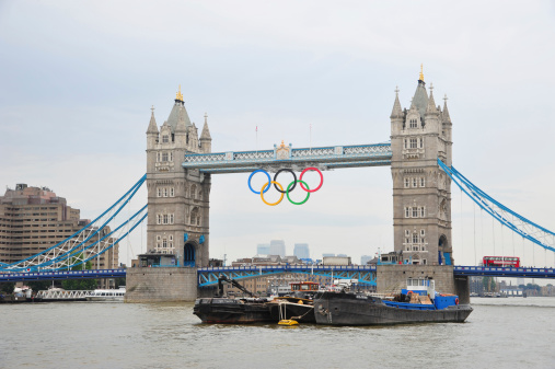 London, UK - August 15, 2012: London - View of Tower Bridge with the giant Olympic Rings suspended to mark the 2012 Summer Olympic games in London. Tour bus drives on London Bridge and boats cruise on river Thames.