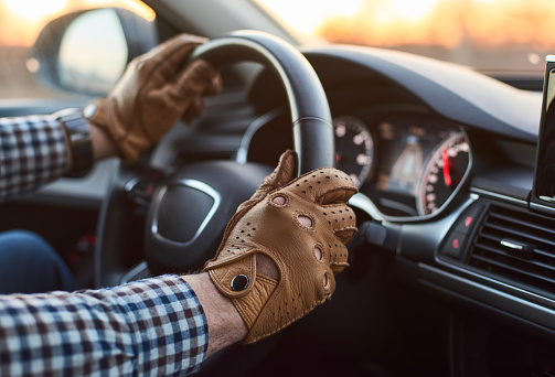 Young person driver in leather gloves travelling in luxury car on sunrise inside view with dashboard, speedometer and gps navigation hands close-up shop