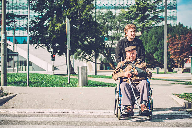 Senior Disabled Man in Wheelchair and Grandson Outdoors, Europe A disabled senior man and his male nurse, grandson, crossing a road in the city centre, Nova Gorica, Slovenia, Europe.  Sunny, cold, autumnal day. Front view. Nikon D800, full frame, XXXL. nova gorica stock pictures, royalty-free photos & images