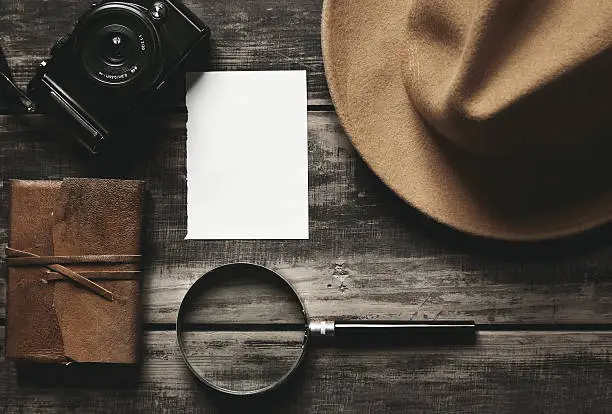 Mysterious detective game concept. Closed notebook in leather cover, sheet of white paper, felt brown hat, portable mirrorless microthird digital photo camera and big vintage magnifier isolated on black aged wood table