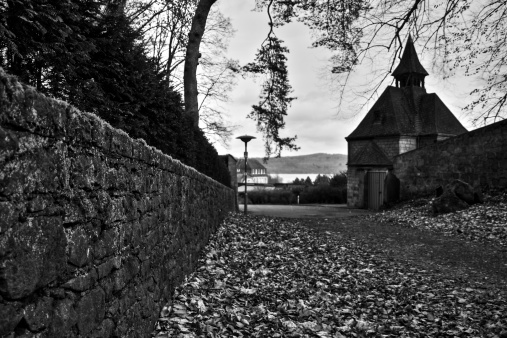 Medieval chapel by a leaf-covered gravel road close to Laacher See in Maria Laach, Germany. An autumn sky sheds a mysterious light on the tranquil scene, giving it a sense of serenity and calm. A lonesome hiking trail, covered in autumn leaves leads toward the lake that is seen in the background of the picture. Old stone walls lead along the trail.