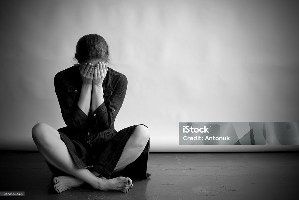 Depressed woman in monochrome Woman is sitting on an old floor with cracks. She is sad and depressed, covering her face with hands. Studio paper  background in behind her. Women Stock Photo