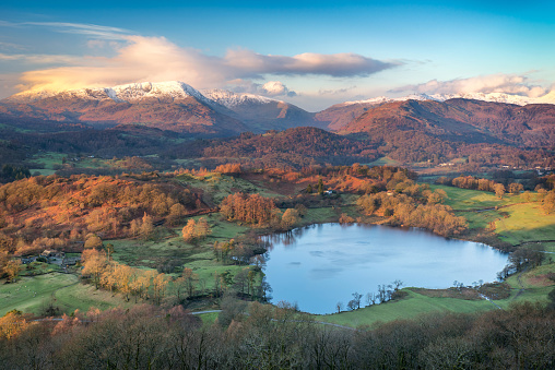 Taken from the top of Loughrigg Fell, a view down to Loughrigg Tarn in the Lake District UK, as early winter sunlight casts a golden red glow across the scene and the clouds, with snow topped fells in the distance.