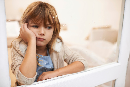 Shot of a bored-looking little girl sitting and looking out a window on a rainy day