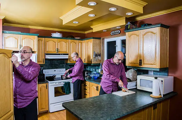 A man so busy in the kitchen that there are three of him cooking, cutting an onion and looking in the kitchen cabinet