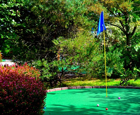 Miniature golf, putt putt golf.  Hole on green turf with flag and golf balls.  Outdoor fun family recreational sporting activity.