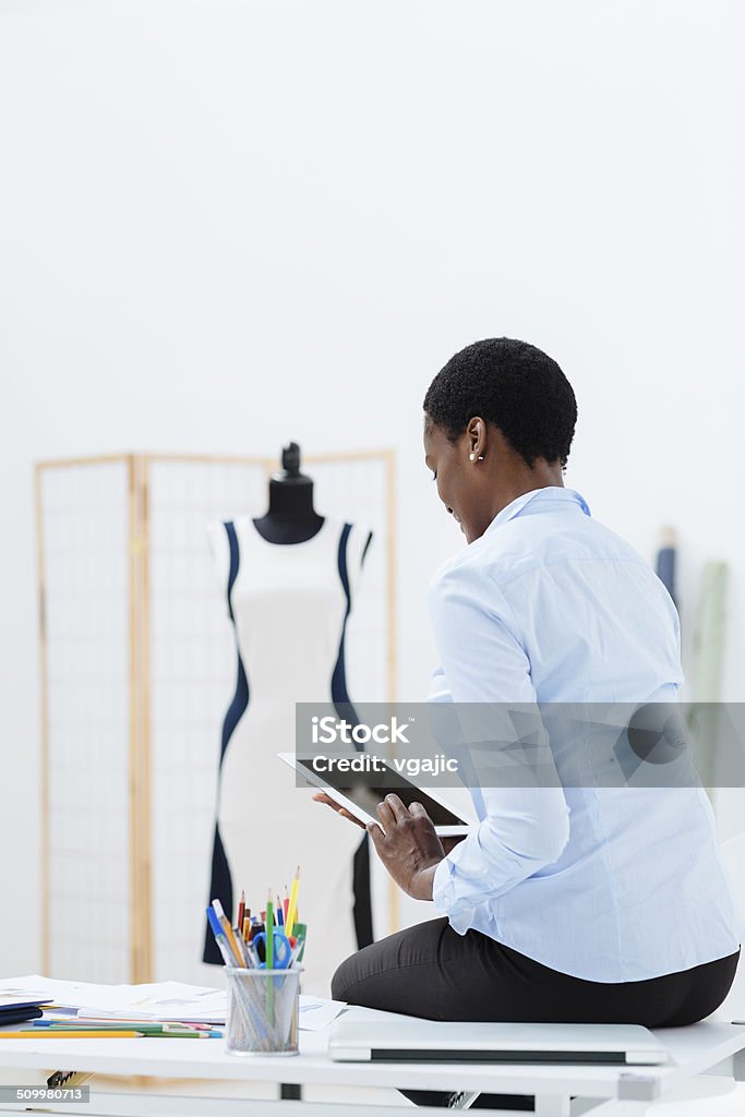 Female fashion designer using digital tablet in office. African woman, fashion designer working in office. Sitting on desk and using digital tablet. 30-39 Years Stock Photo