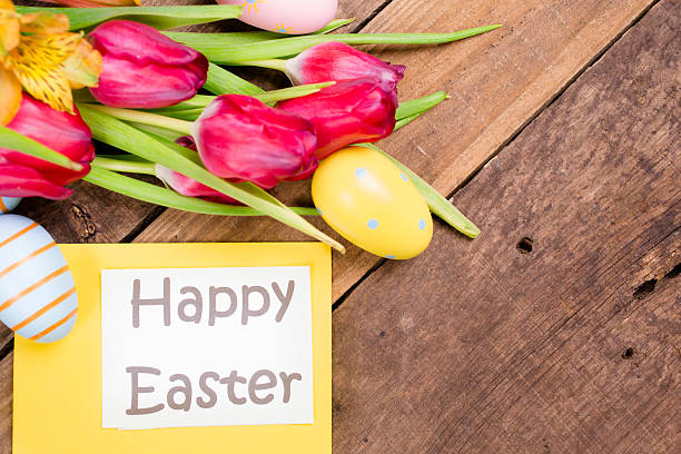 Close-up image of a Happy Easter sign on yellow surrounded by Easter eggs and red tulips all lying on a rustic wooden background. Ready for an Easter Egg Hunt!   Great for Easter and spring themes.  No people. Pastel colors.
