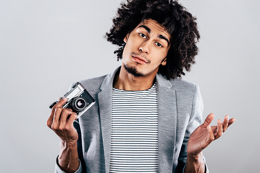 Handsome young African man holding retro styled camera and looking confused while standing against grey background