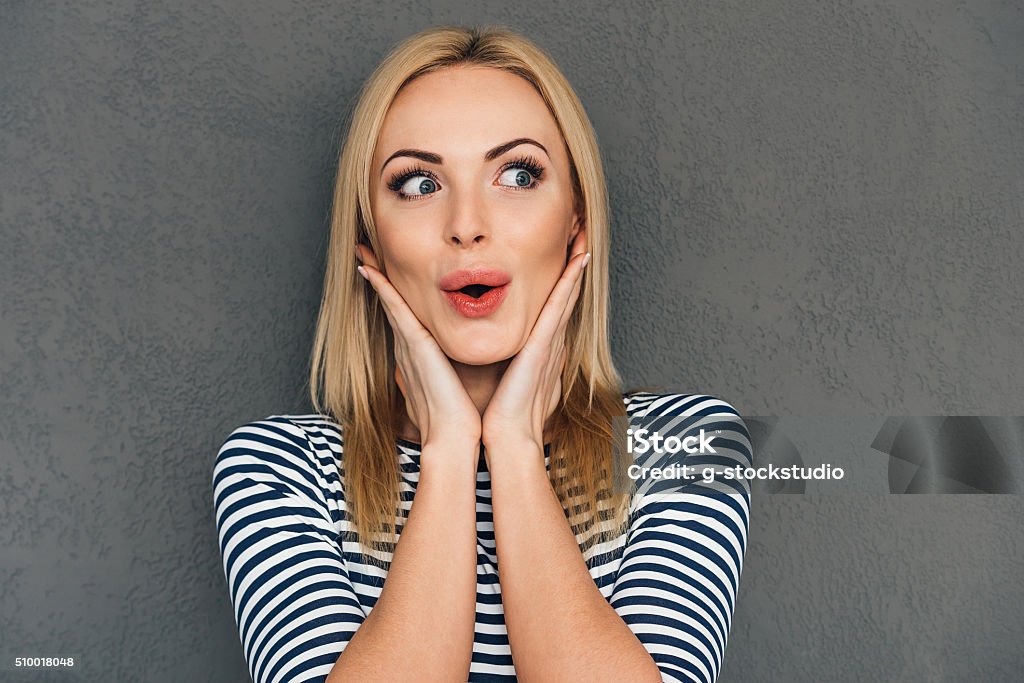 Oh! Beautiful young woman keeping hands on chin and looking surprised while standing against grey background Surprise Stock Photo