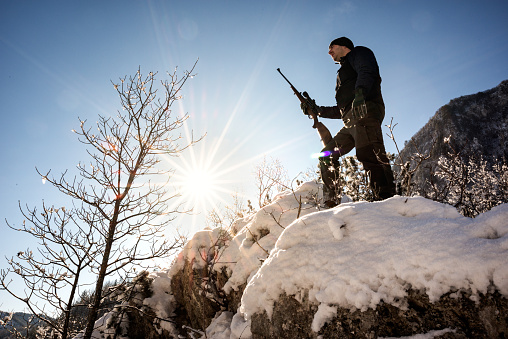 Low angle view of a hunter standing on a snowy hill and holding rifle. He is looking in the distance and sun visible in the sky.