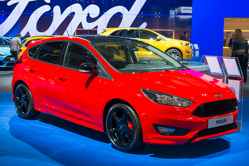 Brussels, Belgium - Januari 12, 2016: Red Ford Focus 5 door hatchback family car. The car is on display during the 2016 Brussels Motor Show. The car is displayed on a motor show stand, with lights reflecting off of the body. There are people looking around and other cars on display in the background.