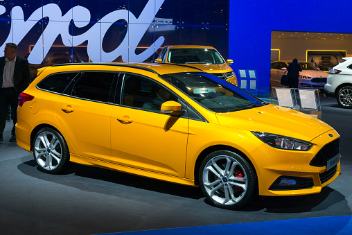 Brussels, Belgium - Januari 12, 2016: Blue Ford Focus Estate station wagon car family car front view. The sporty ST model, features an upgraded version of the 2.3-litre EcoBoost engine. The car is on display during the 2016 Brussels Motor Show. The car is displayed on a motor show stand, with lights reflecting off of the body. There are people looking around and other cars on display in the background.