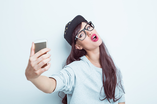 Portrait of beautiful young woman wearing grey jumper, nerd glasses and black baseball cap, taking selfie using a smart phone. Studio shot, white background.
