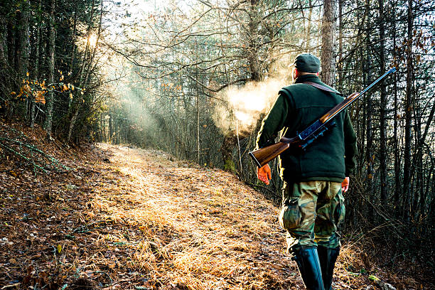Hunter with rifle viewed from behind while walking uphill towards the sunlight that breaches through the trees. Warm breath is visible in front of him.