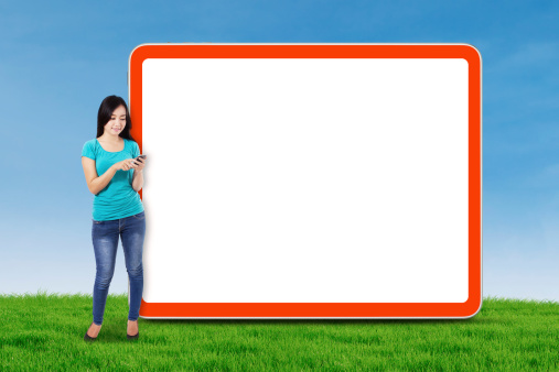 Happy young woman with mobile phone standing next to blank board