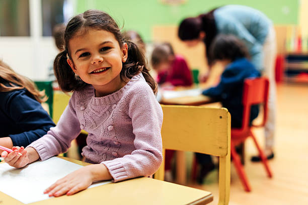 Happy Little Girl in kindergarten Group of children in kindergarten. Showing their imagination and skill and creativity. Focus to little girl sitting, holding pencil in her hand and drawing. Kids around are talking with teacher. Girl looking at camera and smiling. long sleeved recreational pursuit horizontal looking at camera stock pictures, royalty-free photos & images