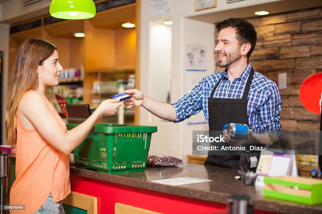 Paying with credit card at a grocery store Profile view of a young woman paying with a credit card to a store clerk in a supermarket Convenience Store Stock Photo