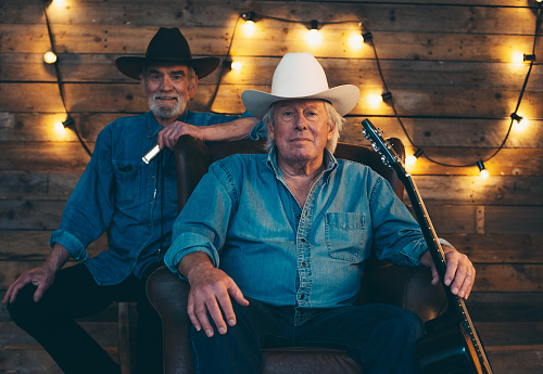 Two senior country and western musicians sitting on chair in front of wooden wall with light bulbs.