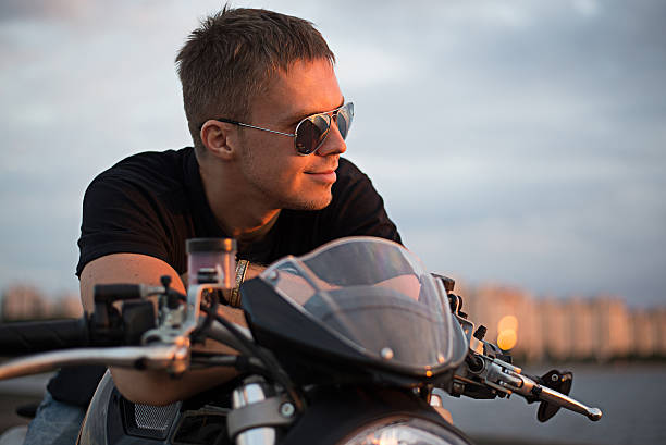 Romantic portrait handsome biker man in sunglasses sits on a bike on a sunset near lake and city