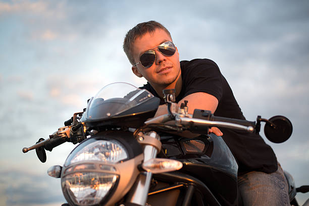 Romantic portrait handsome biker man in sunglasses sits on a bike on a sunset near lake and city
