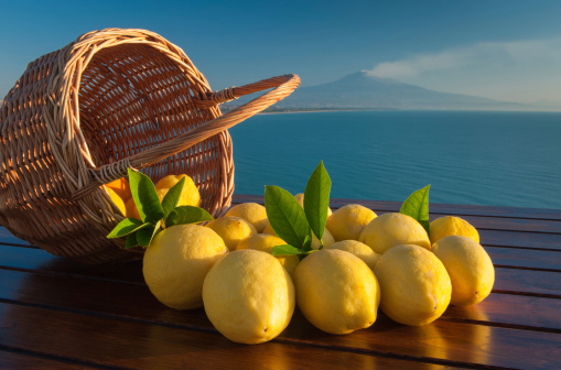 Wicker basket full of  lemons on a wooden table with blue sea and mount Etna in the background